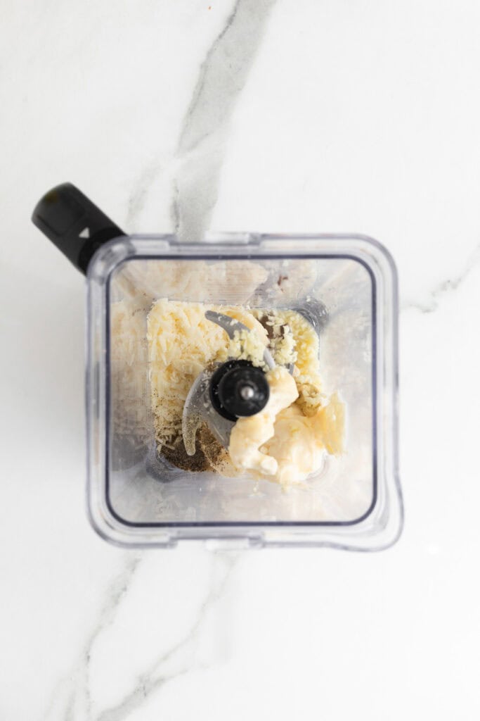 Blender filled with Caesar dressing ingredients like mayo, garlic, and shredded Parmesan