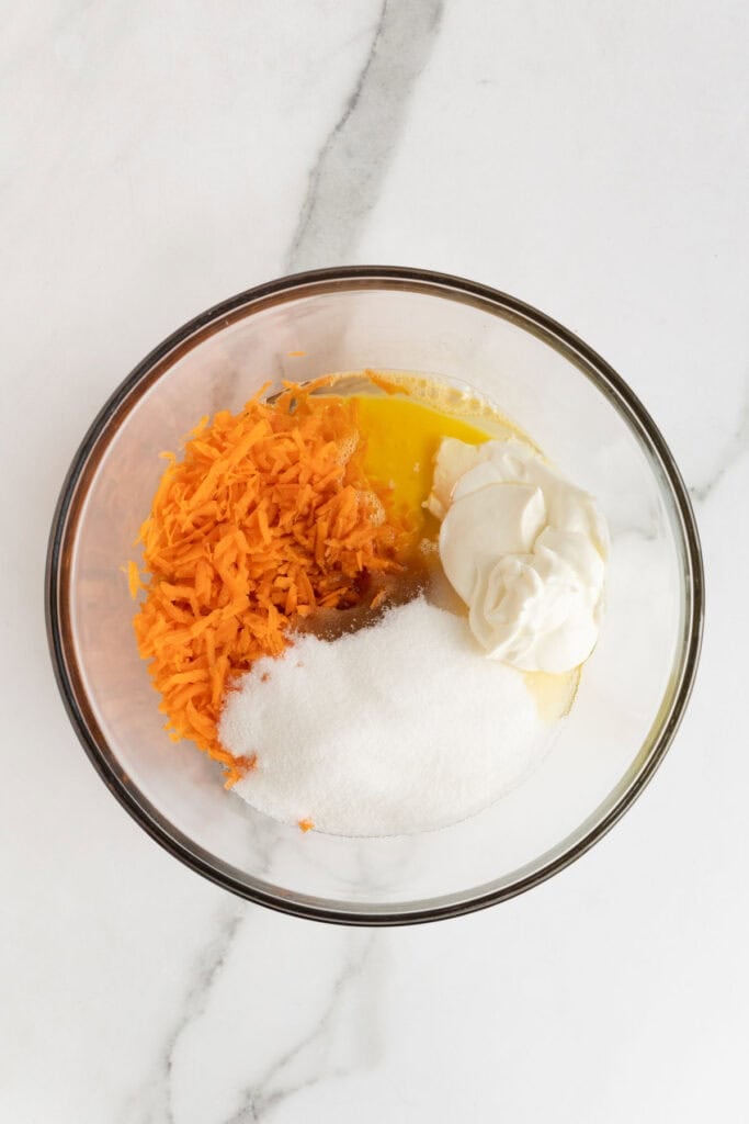 Glass bowl with shredded carrots, sugar, sour cream, eggs, and oil on a marble surface