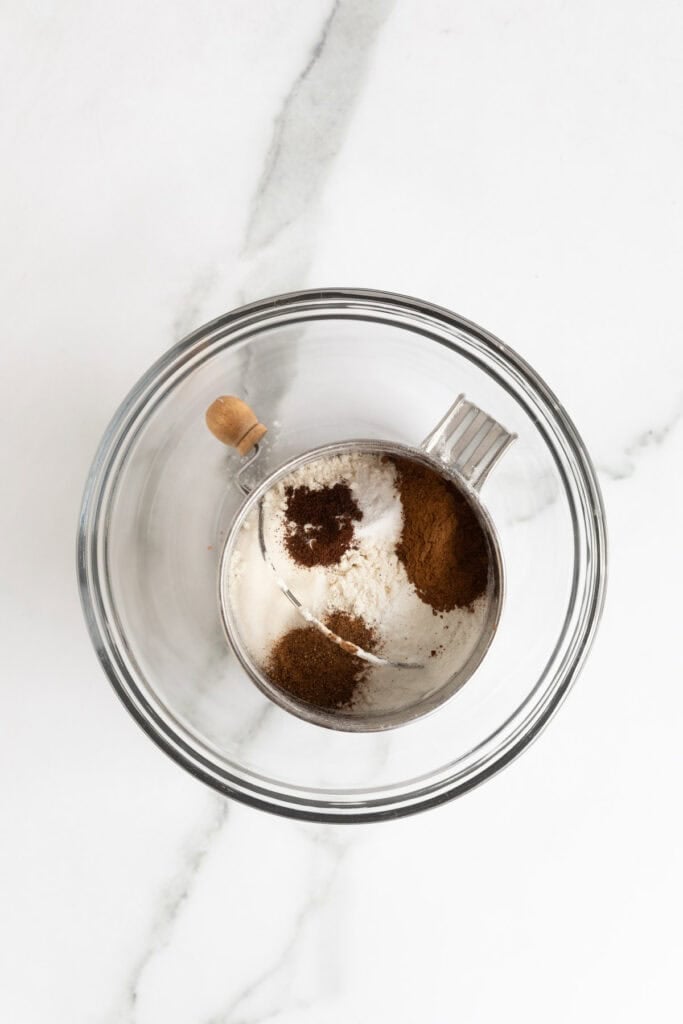 Flour, sugar, baking powder, and spices in a sifter set inside a glass bowl