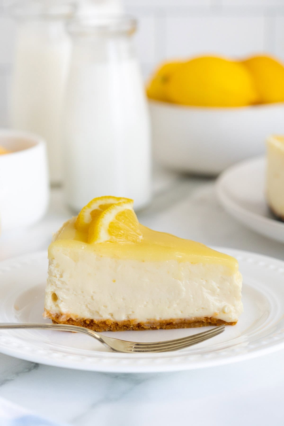 Single slice of lemon cheesecake on a white plate with a fork, topped with lemon slices, with milk bottles and lemons in the background