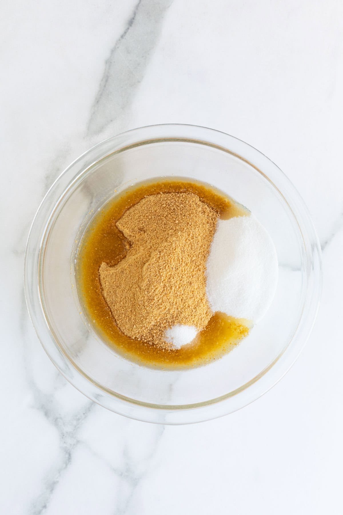 A glass mixing bowl with graham cracker crumbs, sugar, and melted butter before mixing