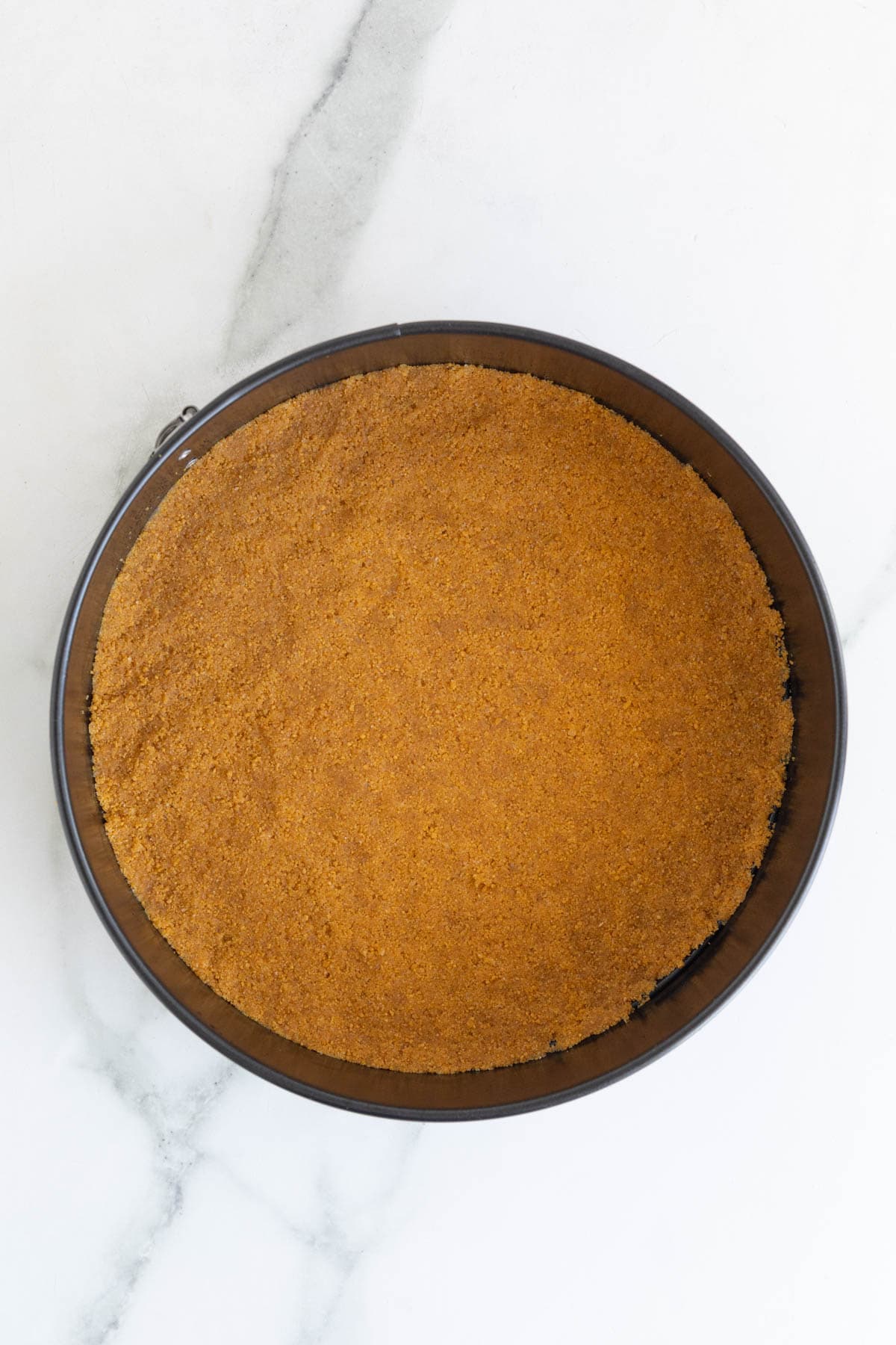 A springform pan filled with a firmly packed graham cracker crust, ready for baking
