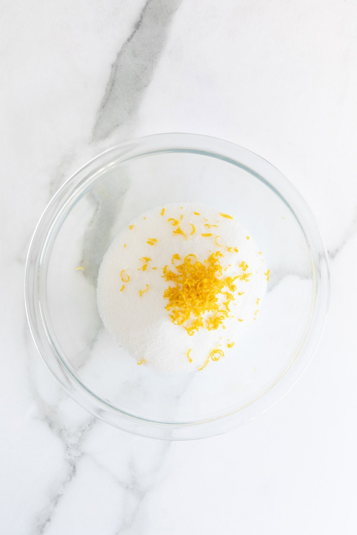 Sugar and lemon zest in a clear bowl on a marble surface