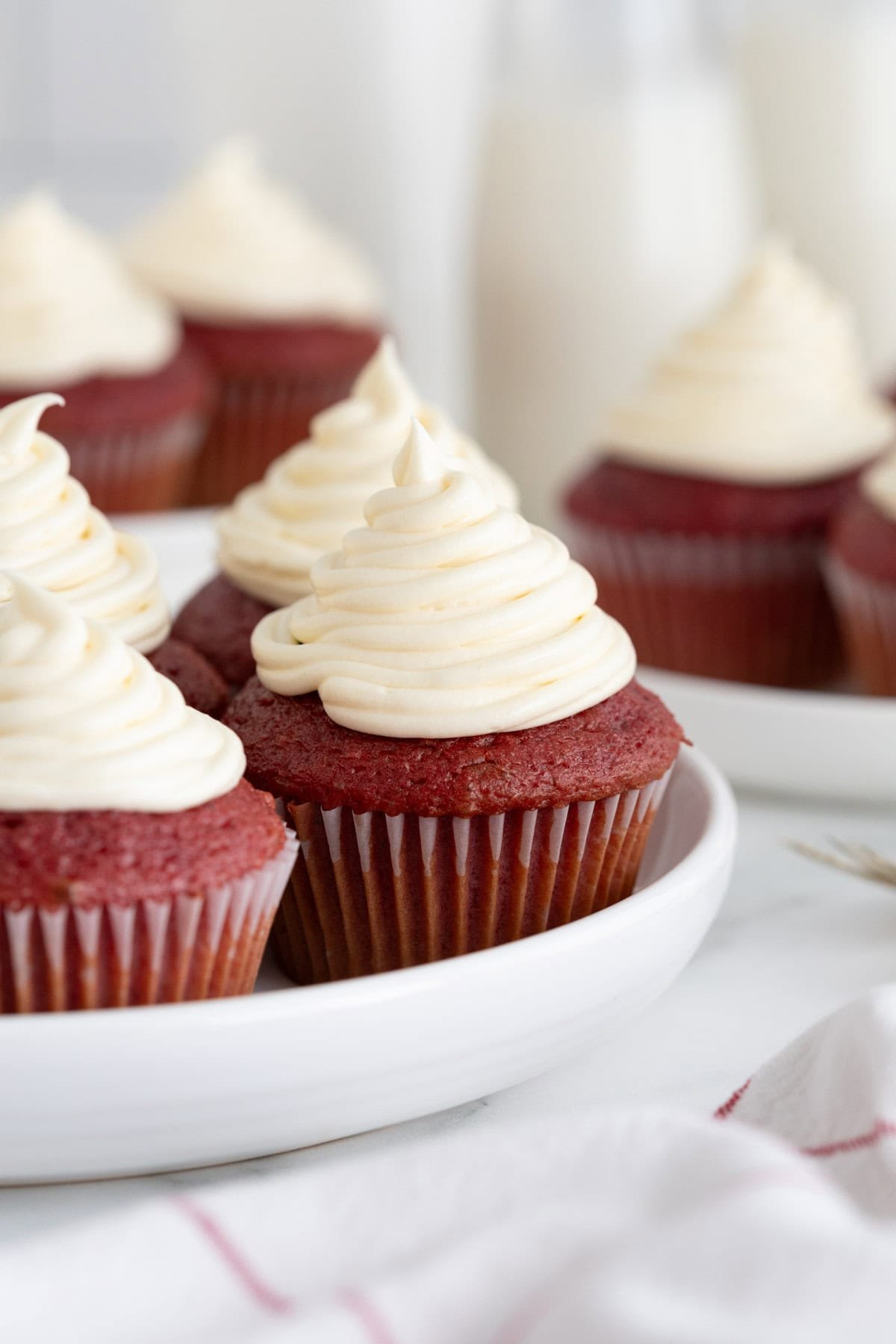 A group of red velvet cupcakes on a white serving plate, each topped with swirled cream cheese frosting