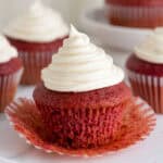 A red velvet cupcake with the wrapper peeled down, showing the moist texture inside, surrounded by more cupcakes