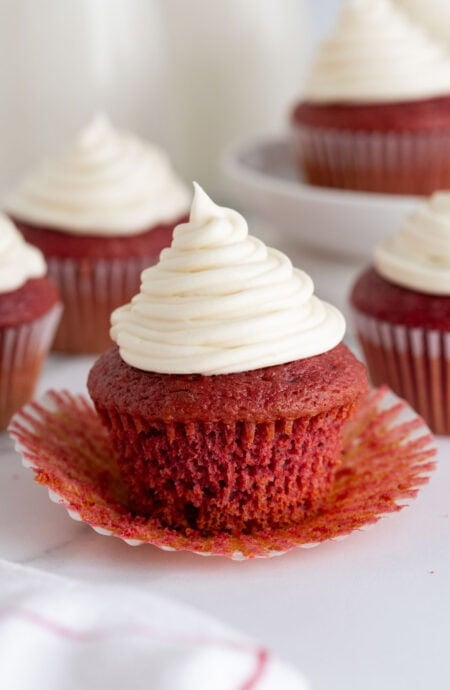 A red velvet cupcake with the wrapper peeled down, showing the moist texture inside, surrounded by more cupcakes