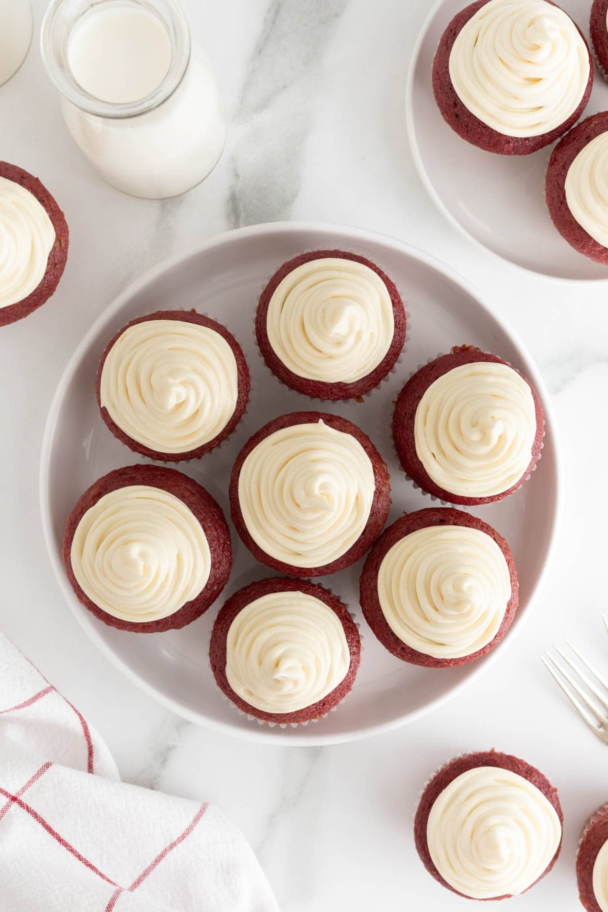 Red velvet cupcakes topped with swirled cream cheese frosting, arranged on a plate with more cupcakes in the background
