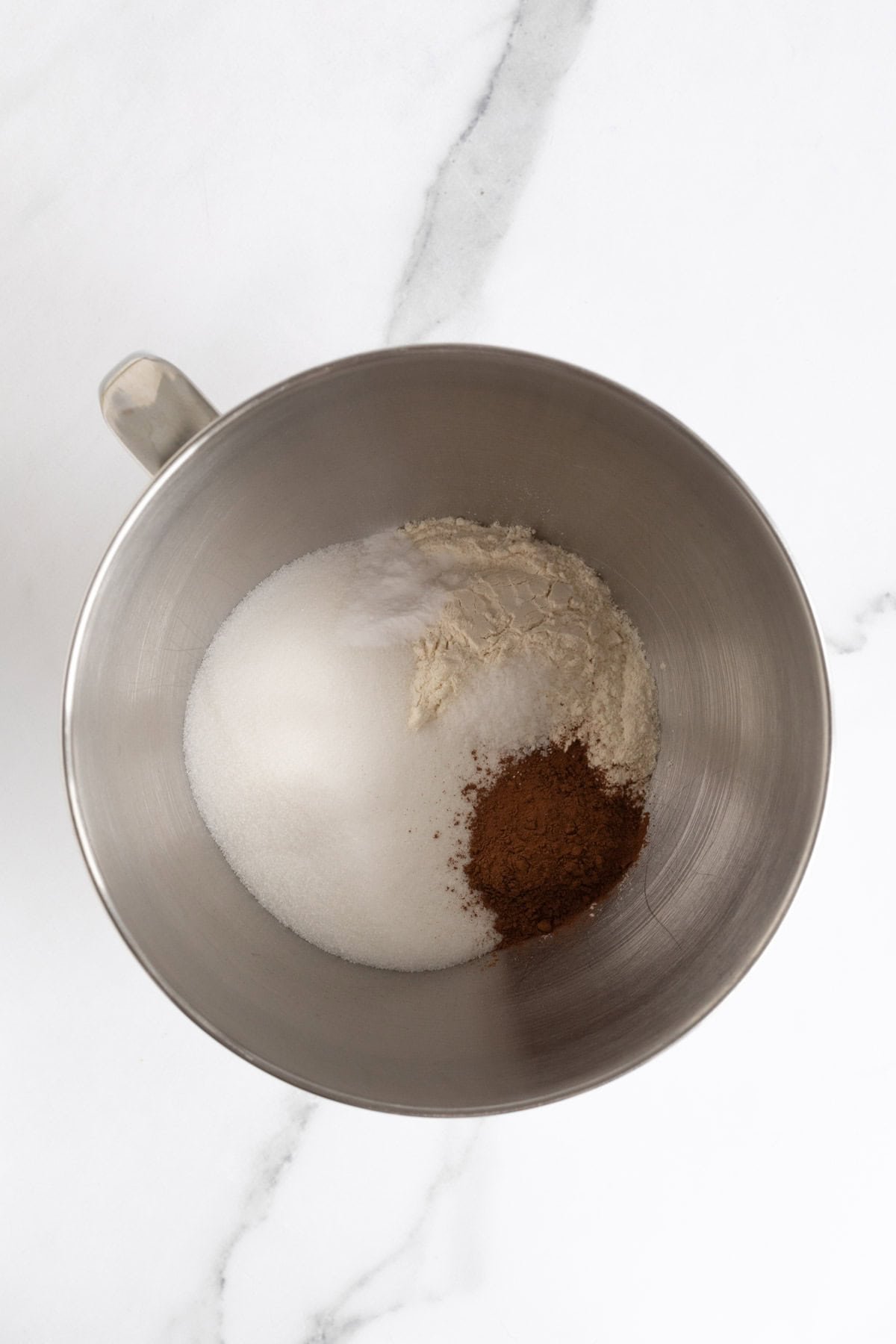 A metal mixing bowl with unmixed dry ingredients including flour, sugar, cocoa powder, and baking soda