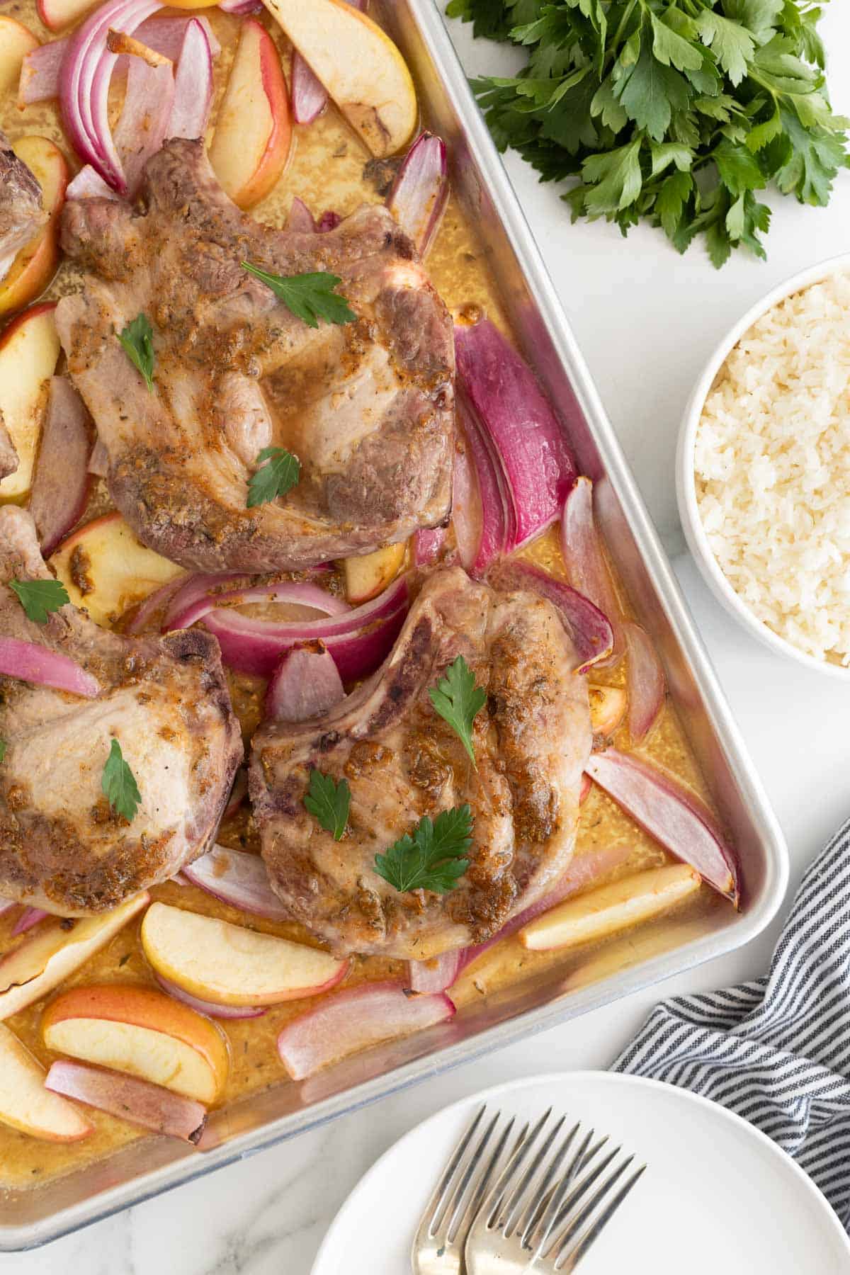 Overhead shot of sheet pan pork chops on a sheet pan with red onions and apple slices, garnished with fresh parsley, with a bowl of white rice next to it