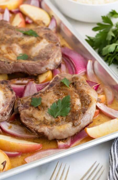 Closeup of sheet pan pork chops with red onions and apples on a sheet pan, topped with parsley leaves