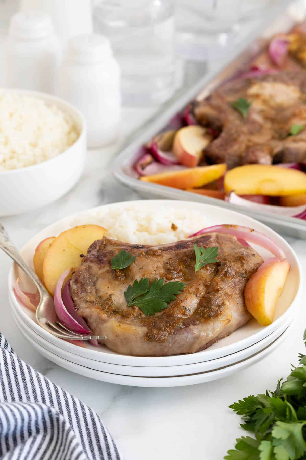Plated sheet pan pork chop served with white rice, apple slices, and red onion, with the full sheet pan in the background