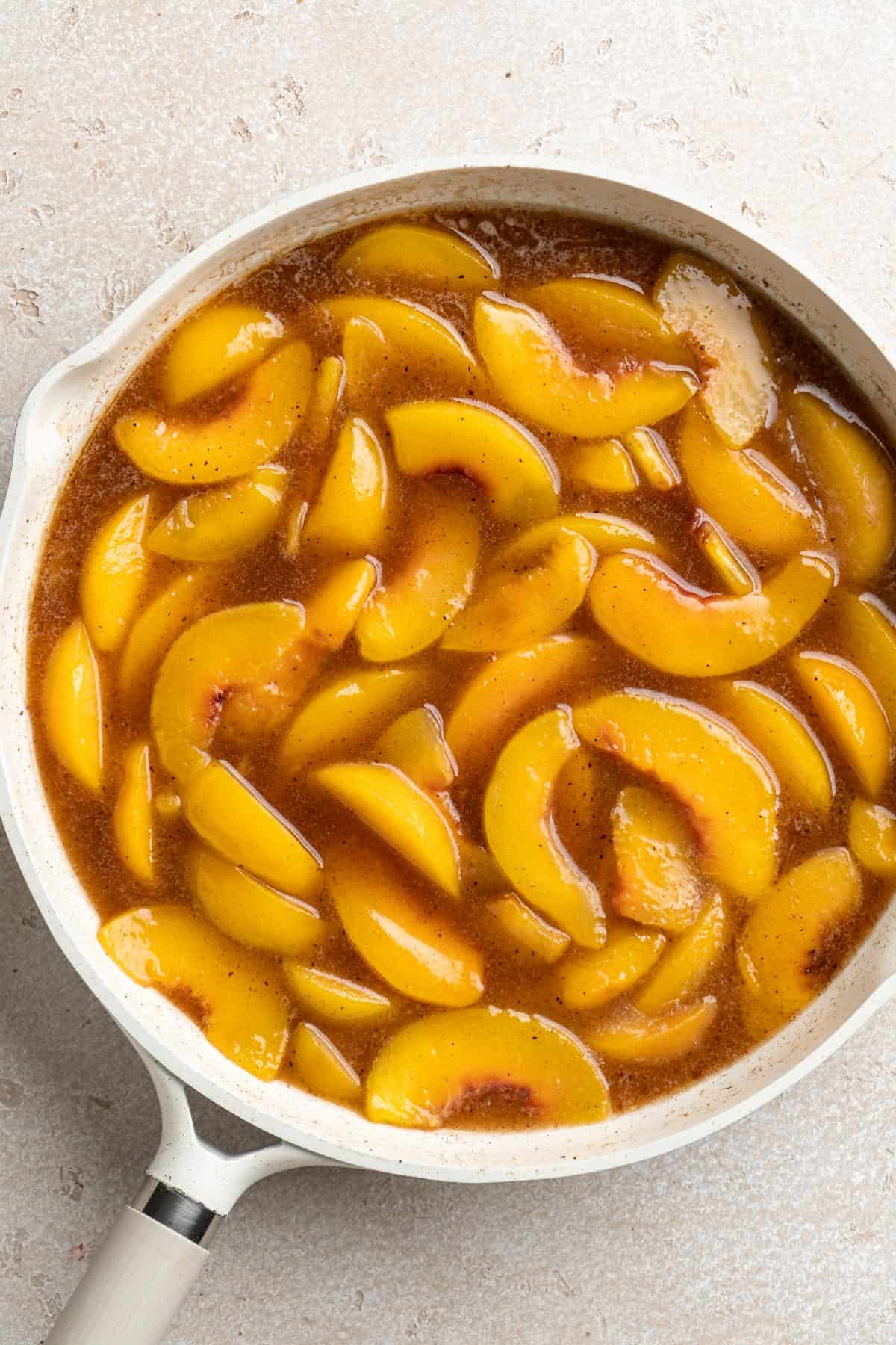 Sliced peaches simmering in a cinnamon-spiced syrup in a white saucepan