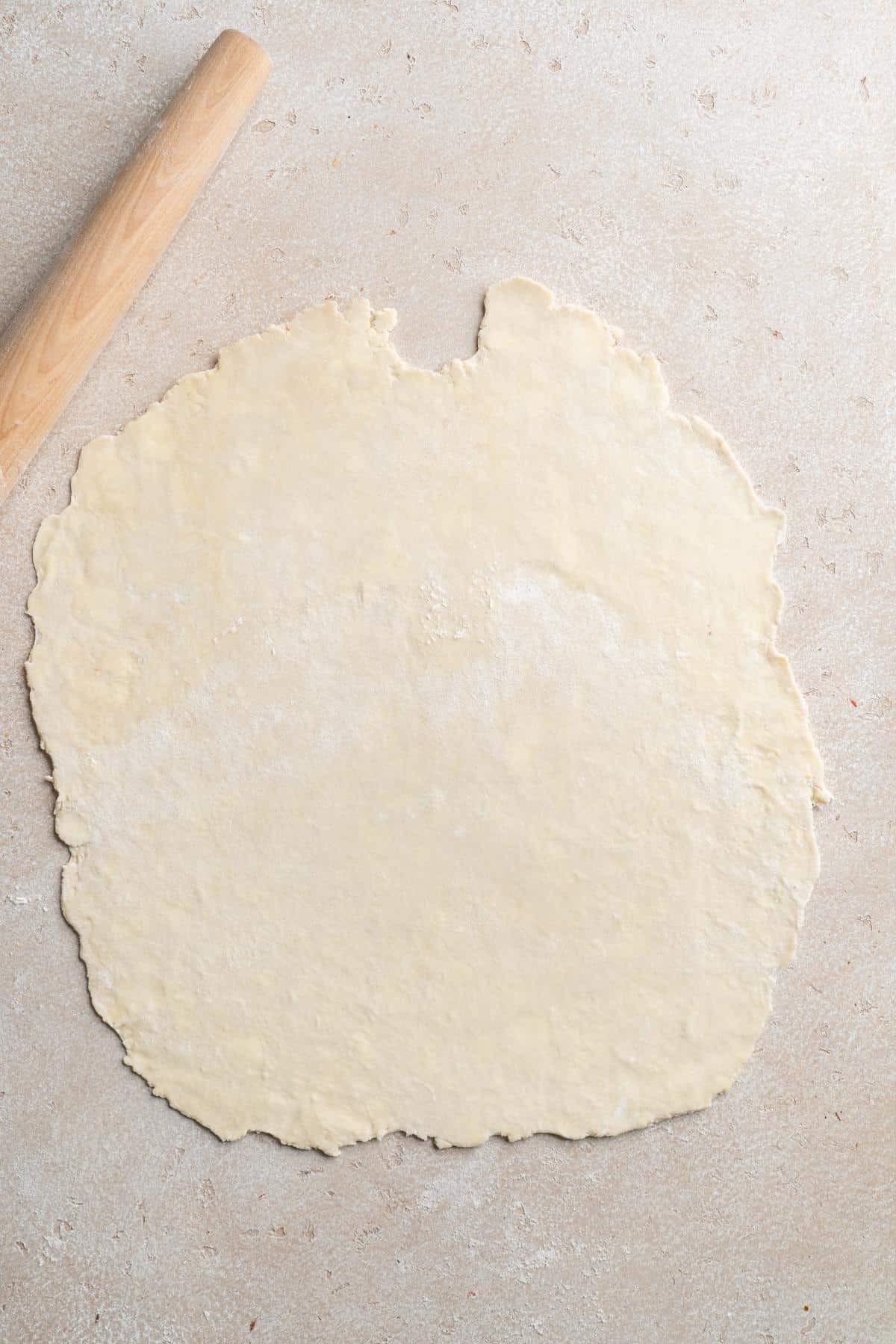 Rolled-out pie crust dough with a wooden rolling pin on a lightly floured beige countertop