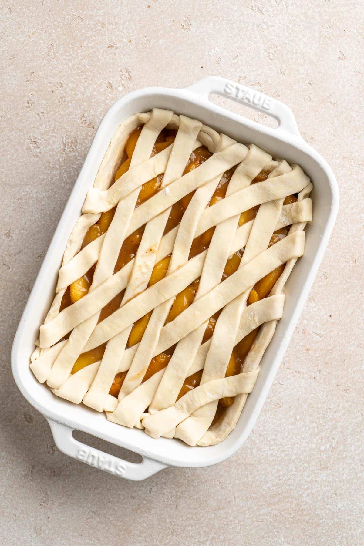Unbaked peach cobbler topped with a lattice pie crust in a white rectangular baking dish