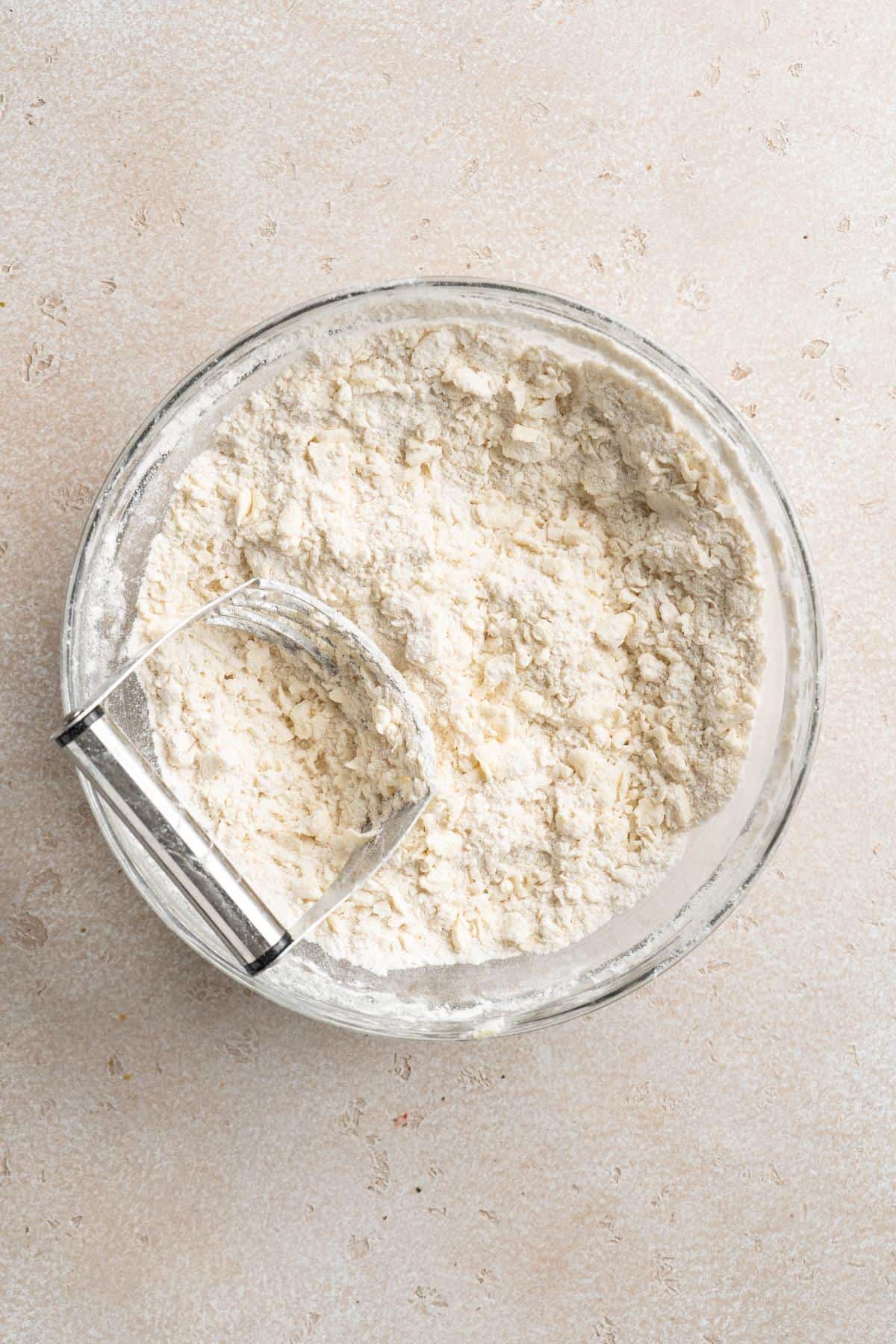 Flour and butter mixture with a pastry cutter in a glass bowl on a beige countertop