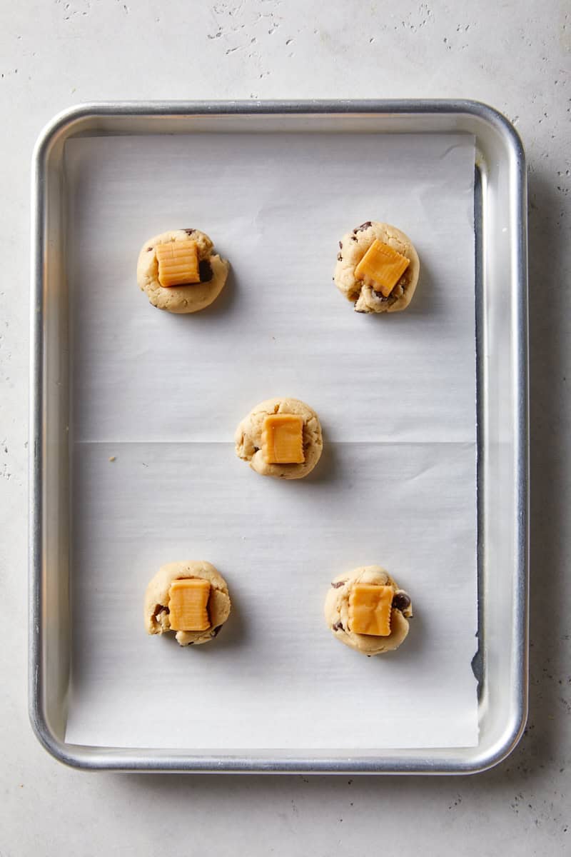 Scoops of chocolate chip cookie dough topped with caramel pieces on a lined baking sheet