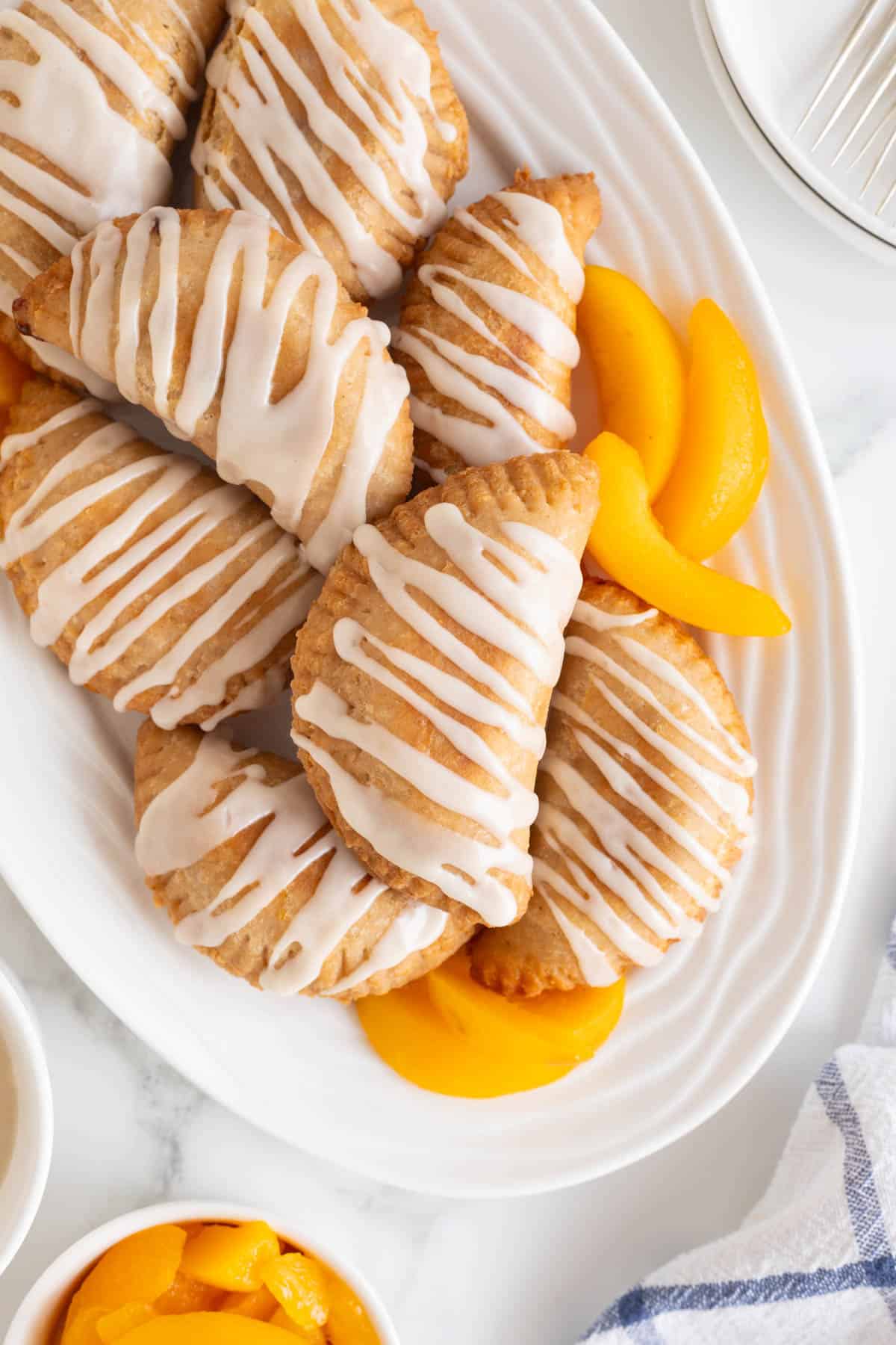 Closeup of golden peach fried pies with icing and peach slices on a white serving platter
