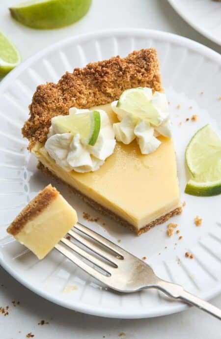 Closeup of a plated slice of key lime pie with whipped cream, lime garnish, and a fork with a bite ready to eat
