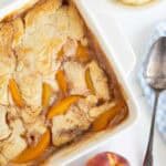 Overhead shot of an easy peach cobbler in a white square dish with a spoon and fresh peaches nearby, and a bowl of ice cream visible in the background, all on a marble surface