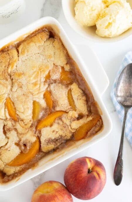 Overhead shot of an easy peach cobbler in a white square dish with a spoon and fresh peaches nearby, and a bowl of ice cream visible in the background, all on a marble surface