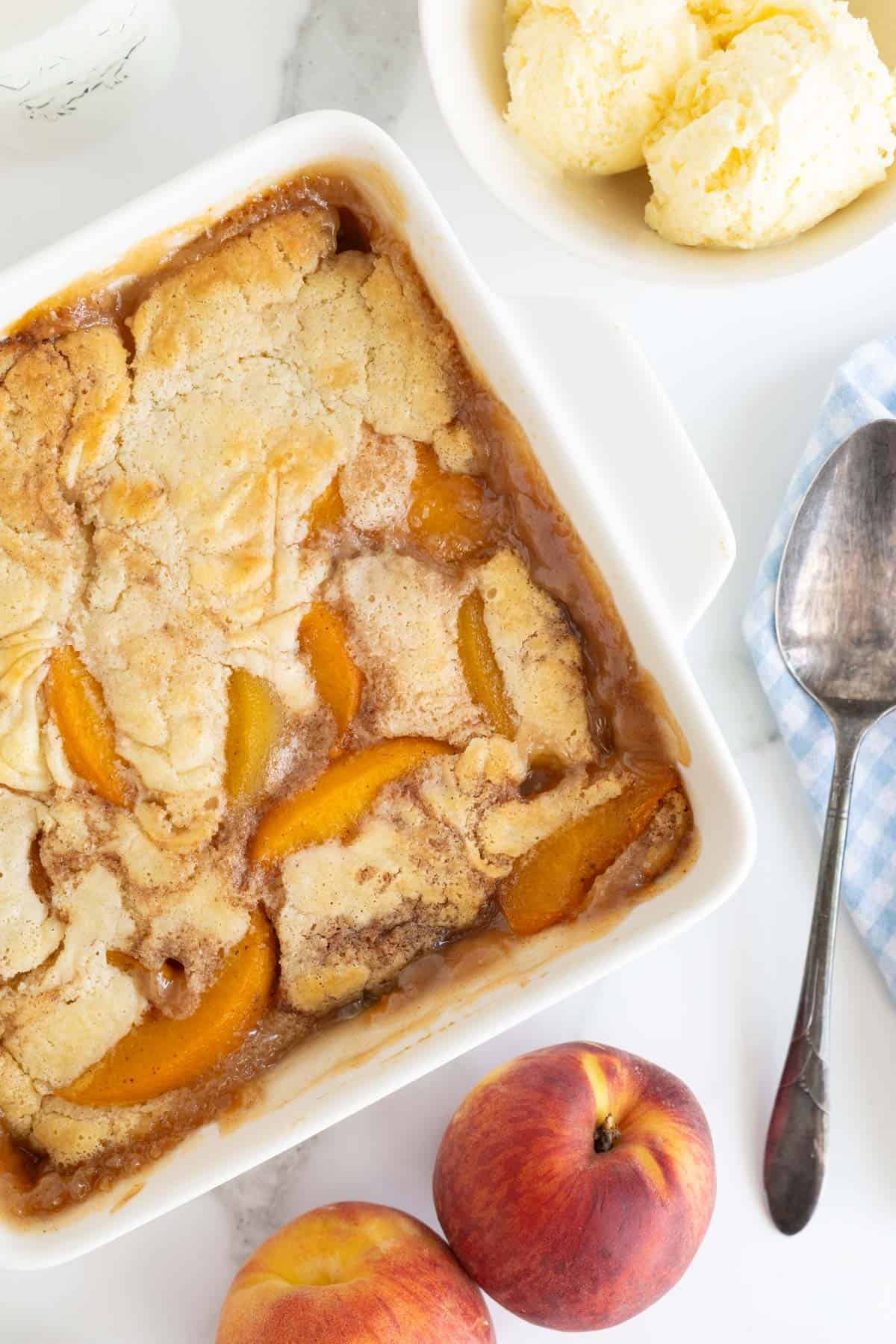 Overhead shot of an easy peach cobbler in a white square dish with a spoon and fresh peaches nearby, and a bowl of ice cream visible in the background, all on a marble surface