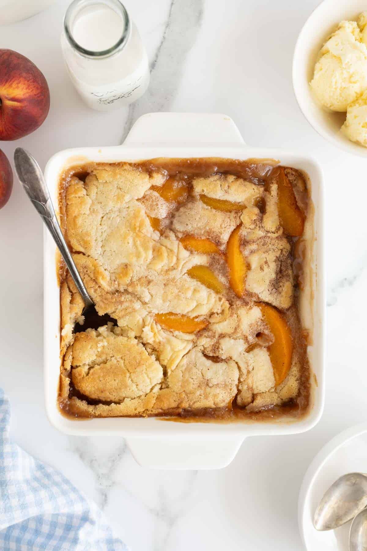 A baked peach cobbler in a white square dish with a spoon, two fresh peaches, a bottle of milk, and a bowl of ice cream on a marble surface