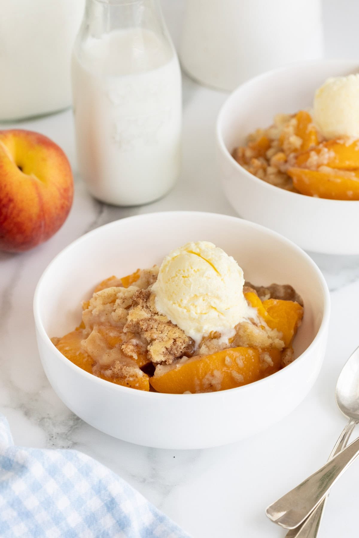 Two white bowls of easy peach cobbler topped with vanilla ice cream, with fresh peaches and milk bottles in the background on a marble counter