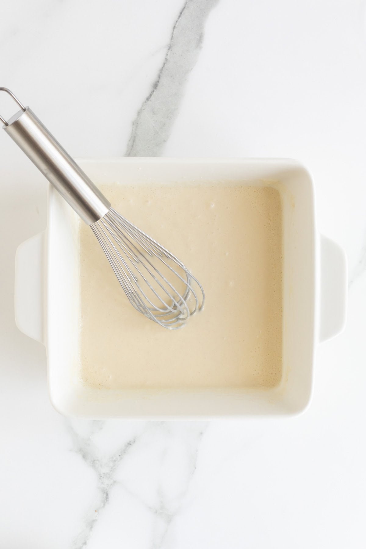 A white square baking dish on a marble counter with a light-colored batter with a whisk resting in it
