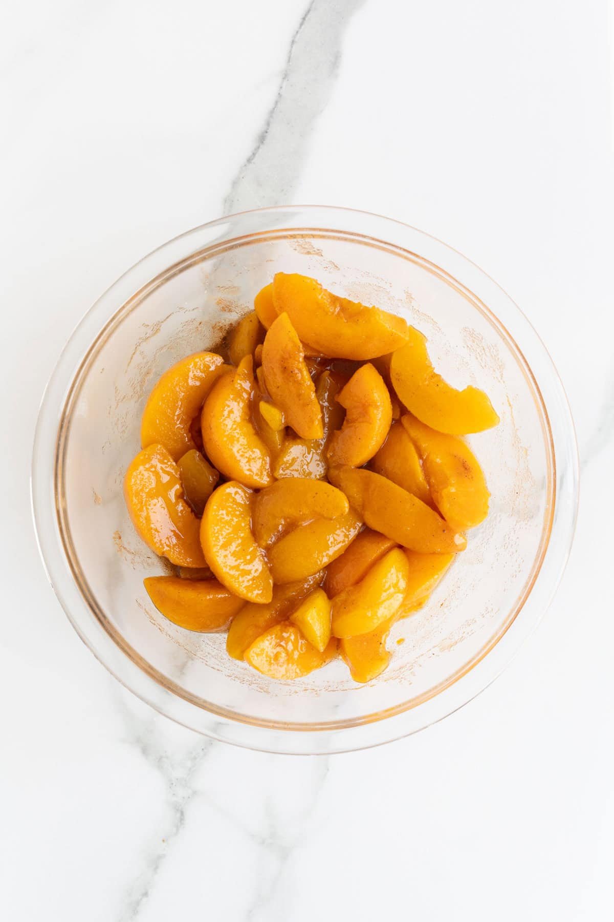 A clear glass bowl on a marble counter with sliced peaches coated in a syrupy, spiced mixture