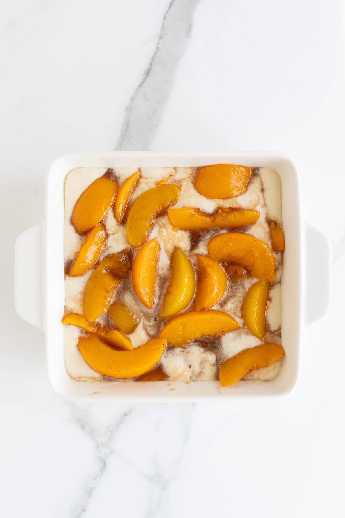 A white square baking dish on a marble counter with a layer of batter topped with sliced peaches