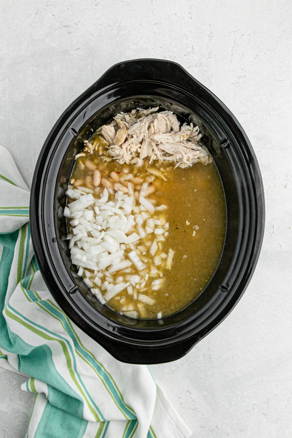 Slow cooker filled with broth, shredded chicken, white beans, and chopped onions before cooking