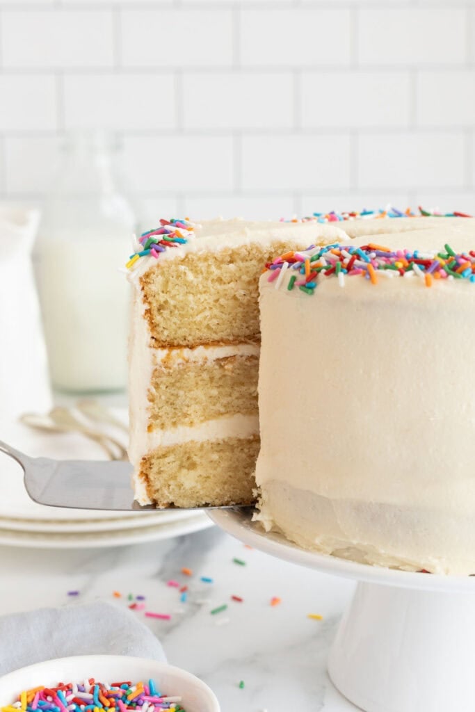 A slice of vanilla cake recipe being removed from full cake on a cake stand
