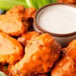 Closeup of buffalo wings coated in sauce with ranch dip on the side and celery sticks in the background