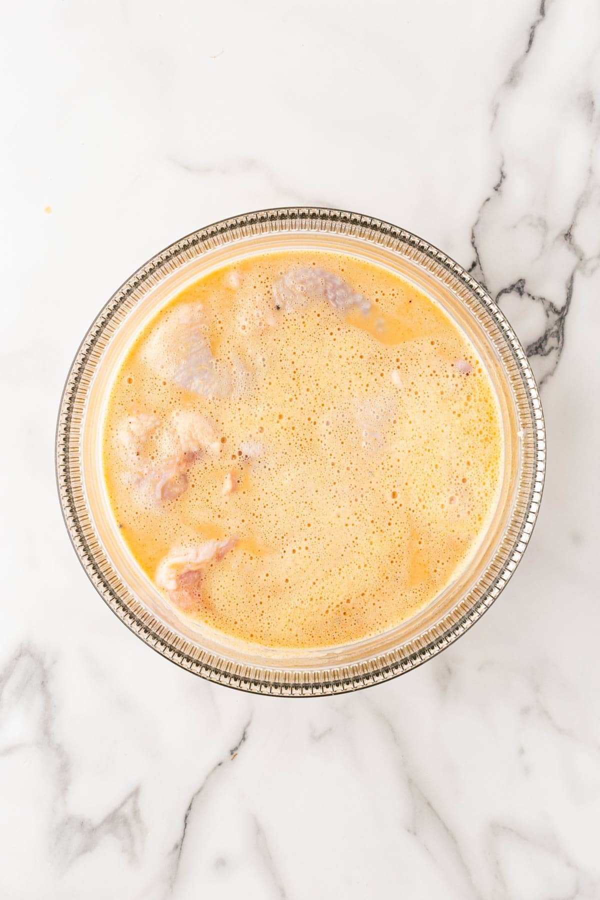 Raw chicken pieces soaking in a seasoned buttermilk marinade in a clear glass bowl