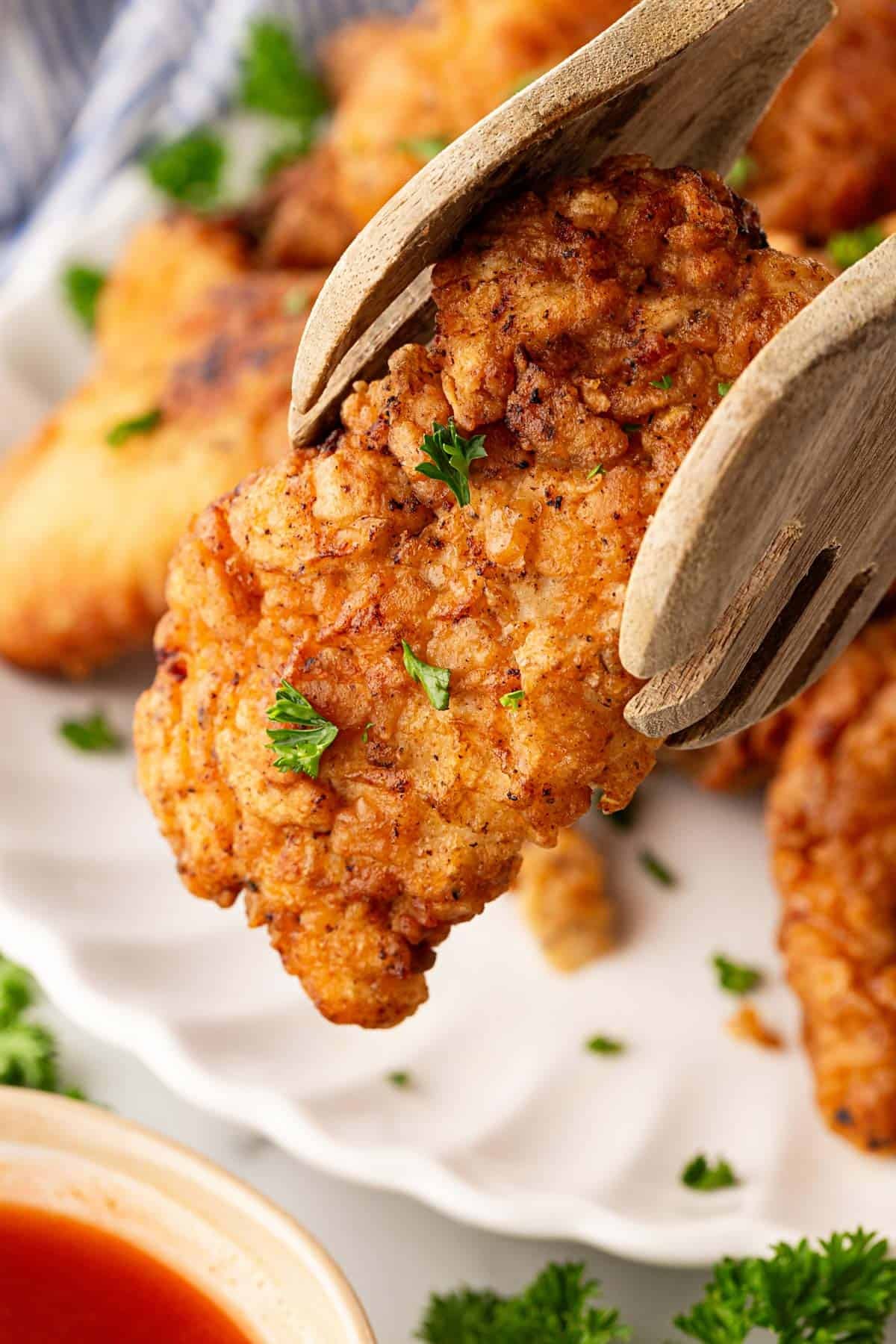 Wooden tongs holding a crispy piece of buttermilk fried chicken above the serving platter