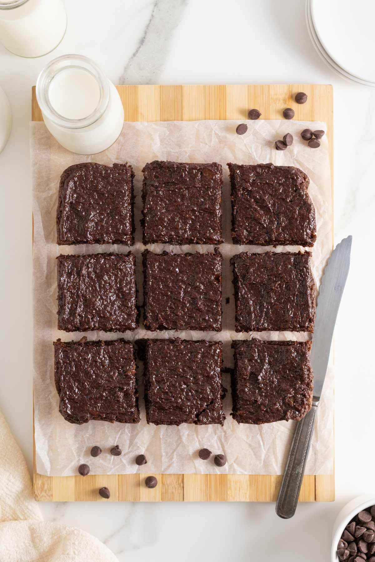 Nine frosted sweet potato brownies arranged in a 3x3 grid on parchment paper over a wooden board, with a knife and chocolate chips nearby