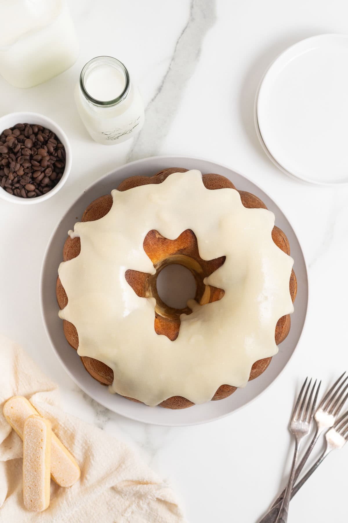 Overhead view of a bundt cake covered in creamy glaze, surrounded by forks, ladyfingers, and milk bottles