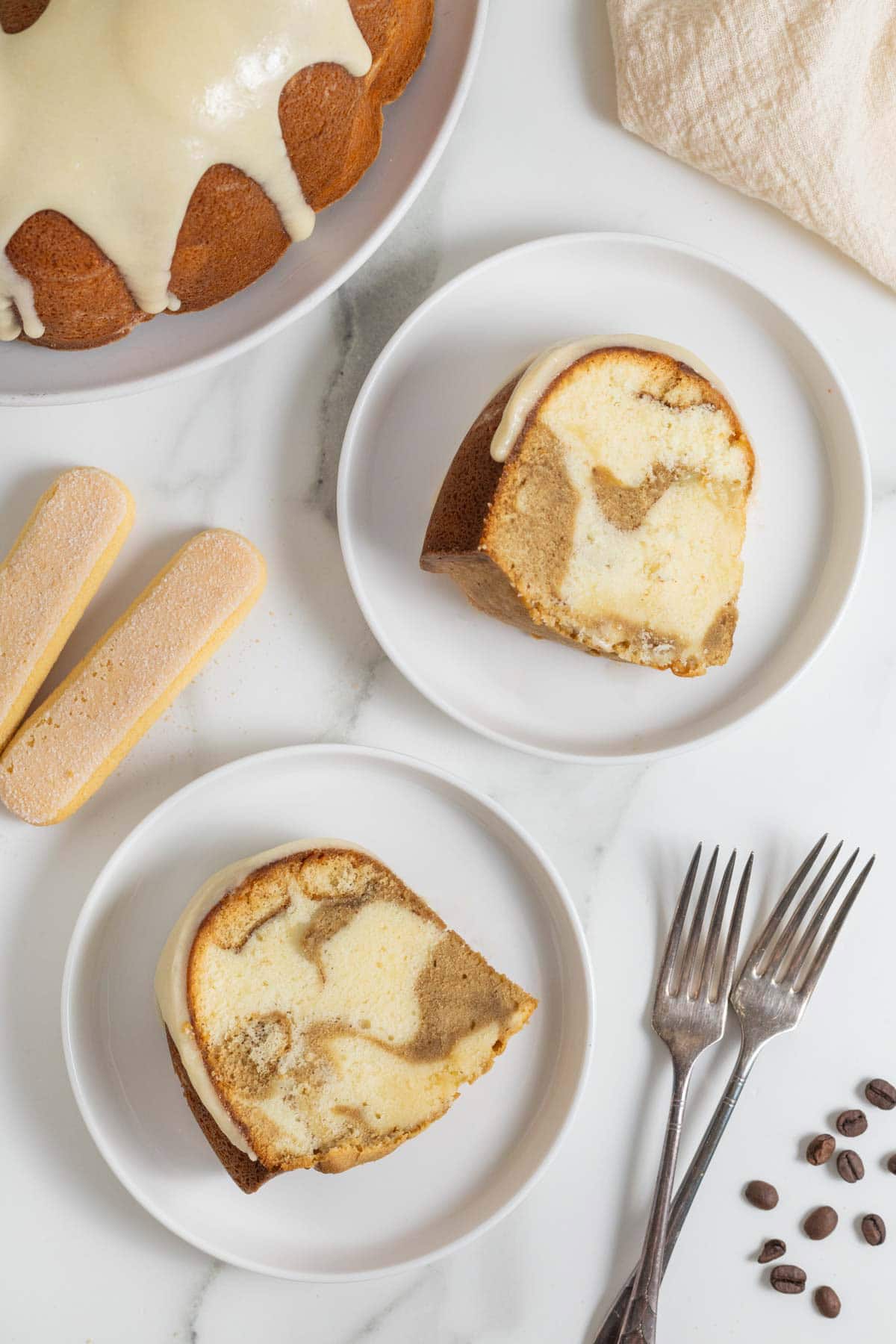 Overhead view of two slices of tiramisu pound cake on plates next to coffee beans, forks, and ladyfingers