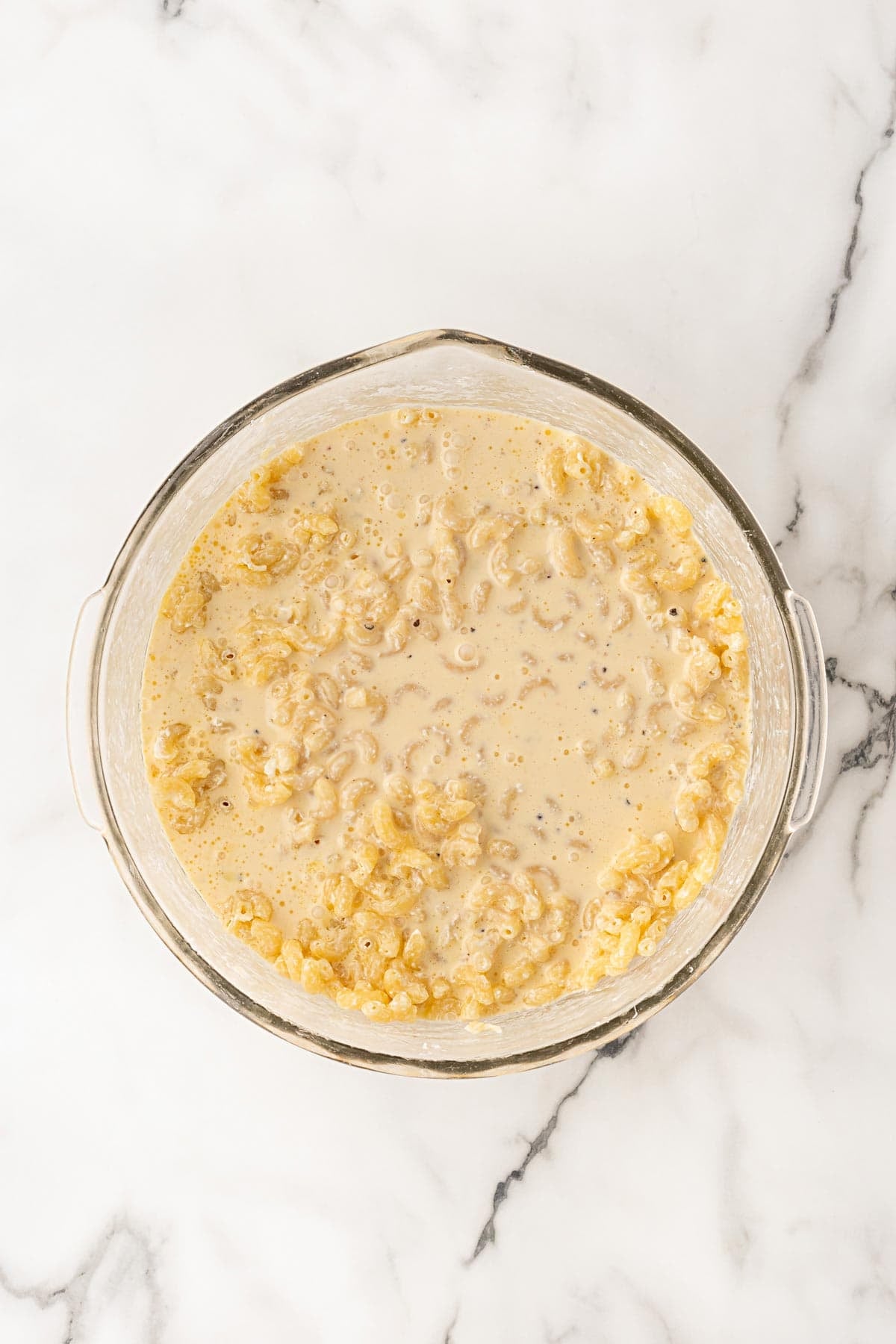 Milk and egg mixture poured over cooked elbow macaroni in a glass bowl