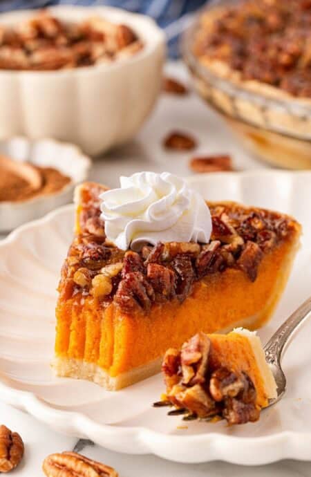 Slice of sweet potato pecan pie topped with whipped cream on a white plate, with a fork holding a bite and pecans scattered nearby