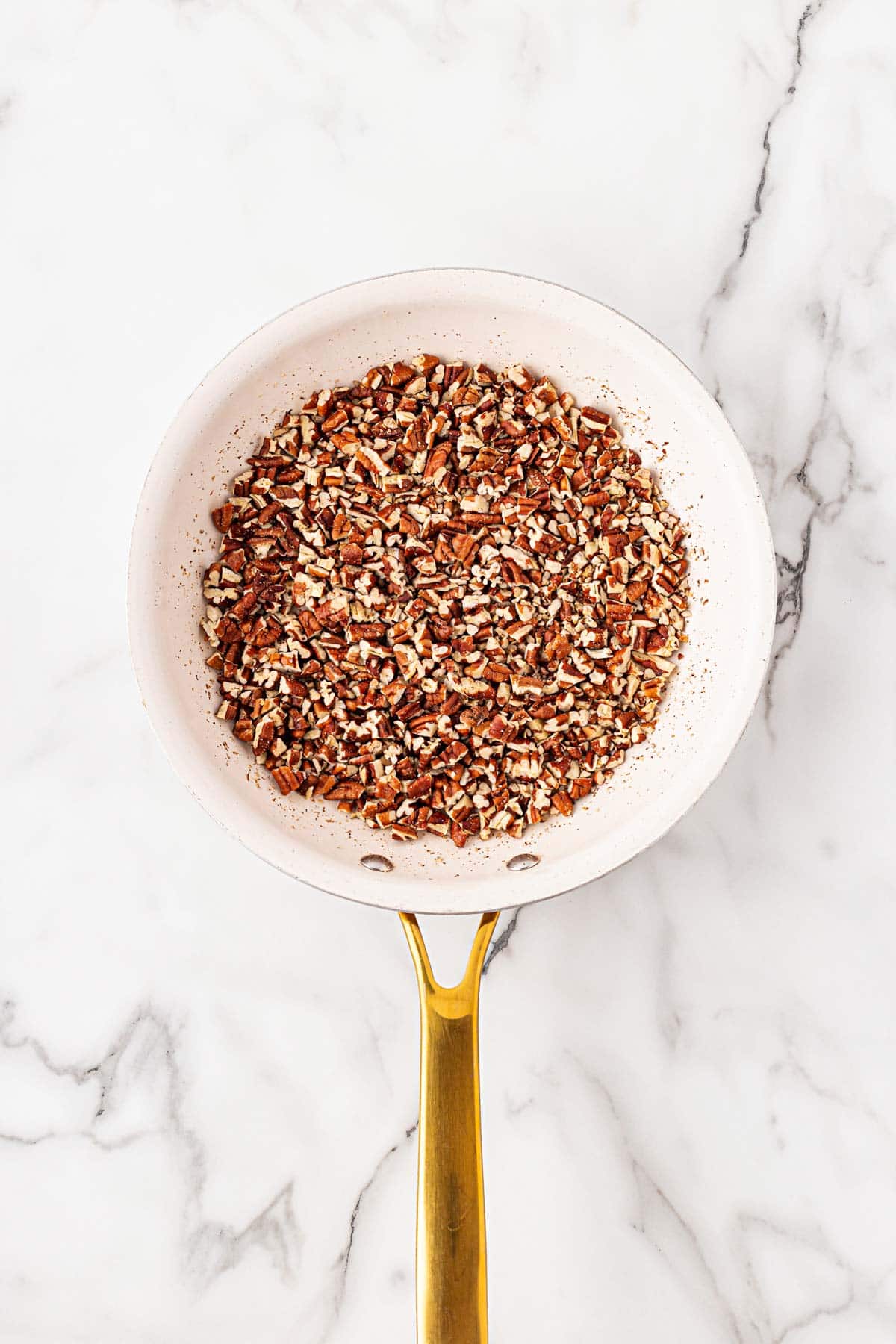 Overhead shot of chopped pecans in a white skillet being lightly toasted on a marble surface
