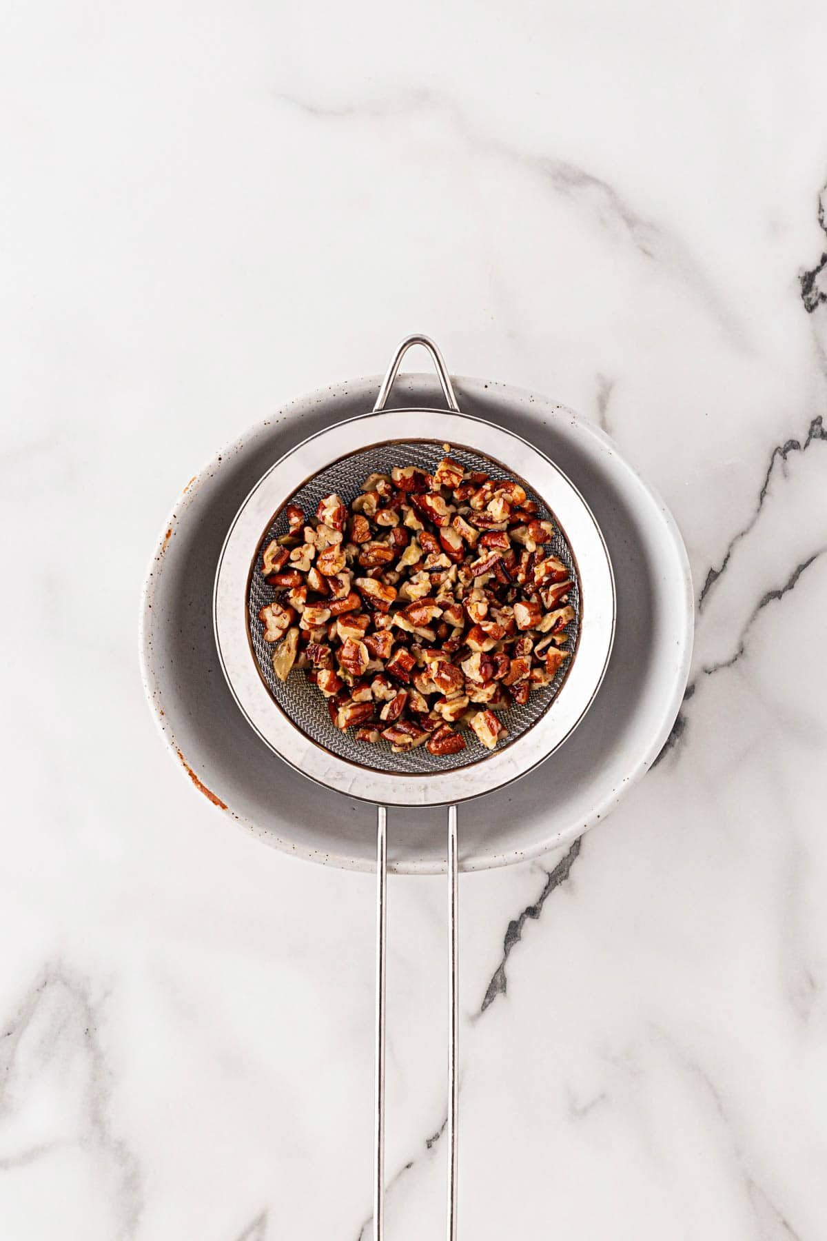 Small mesh strainer with toasted buttered pecans resting over a bowl to drain excess butter