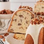 Slice of butter pecan pound cake being lifted from the whole cake, showing its moist, dense crumb filled with pecans and creamy glaze on top