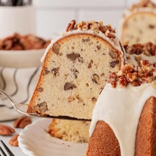 Slice of butter pecan pound cake being lifted from the whole cake, showing its moist, dense crumb filled with pecans and creamy glaze on top