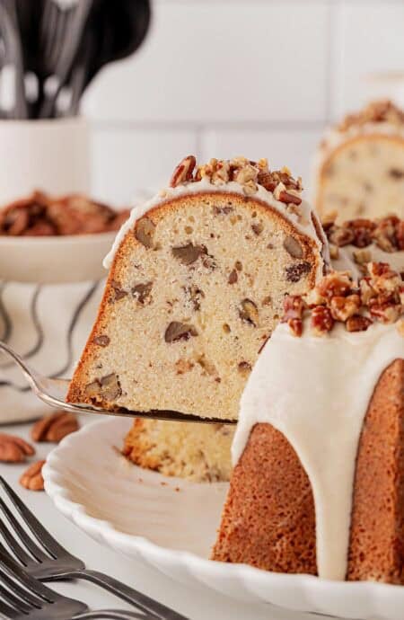 Slice of butter pecan pound cake being lifted from the whole cake, showing its moist, dense crumb filled with pecans and creamy glaze on top