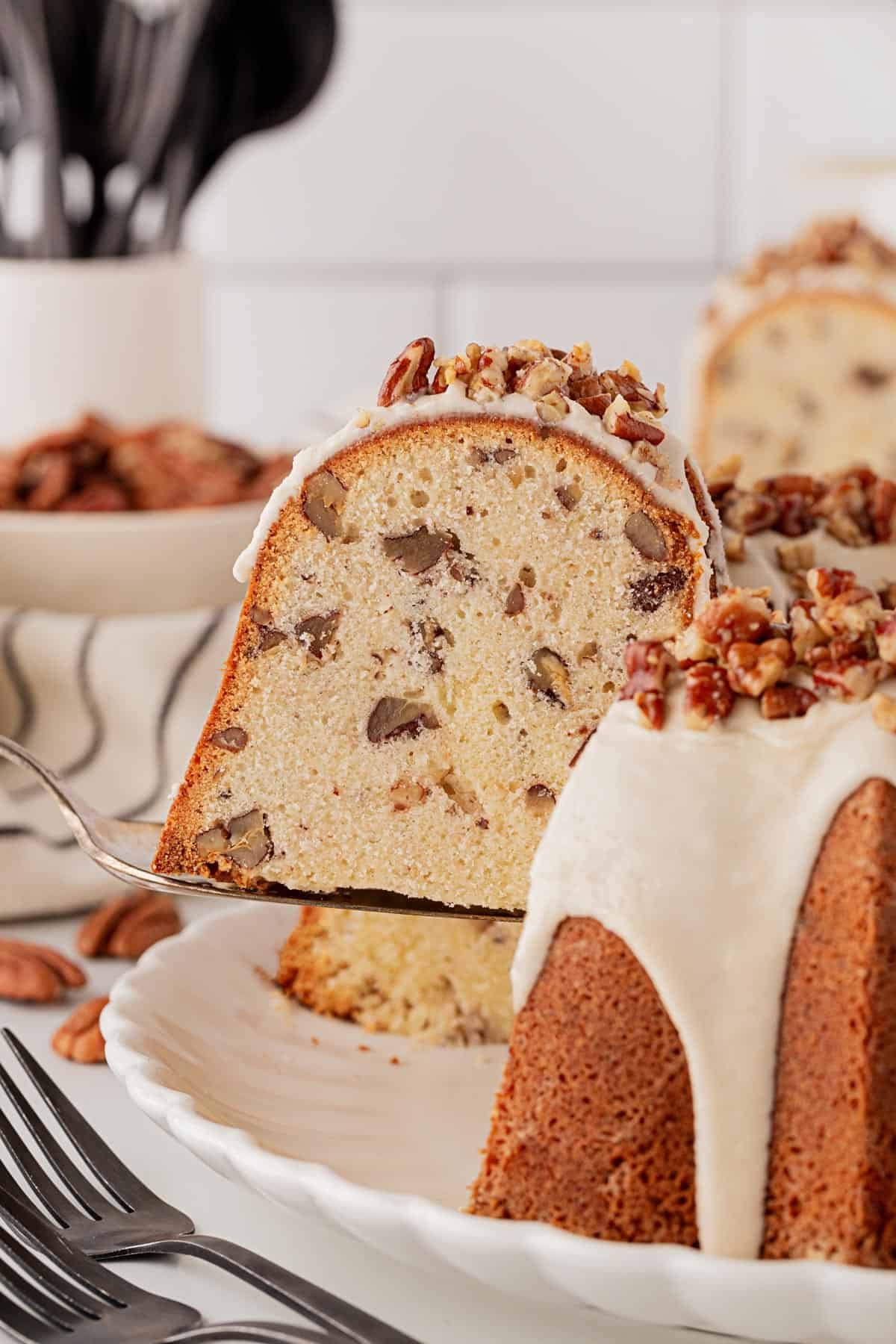 Slice of butter pecan pound cake being lifted from the whole cake, showing its moist, dense crumb filled with pecans and creamy glaze on top
