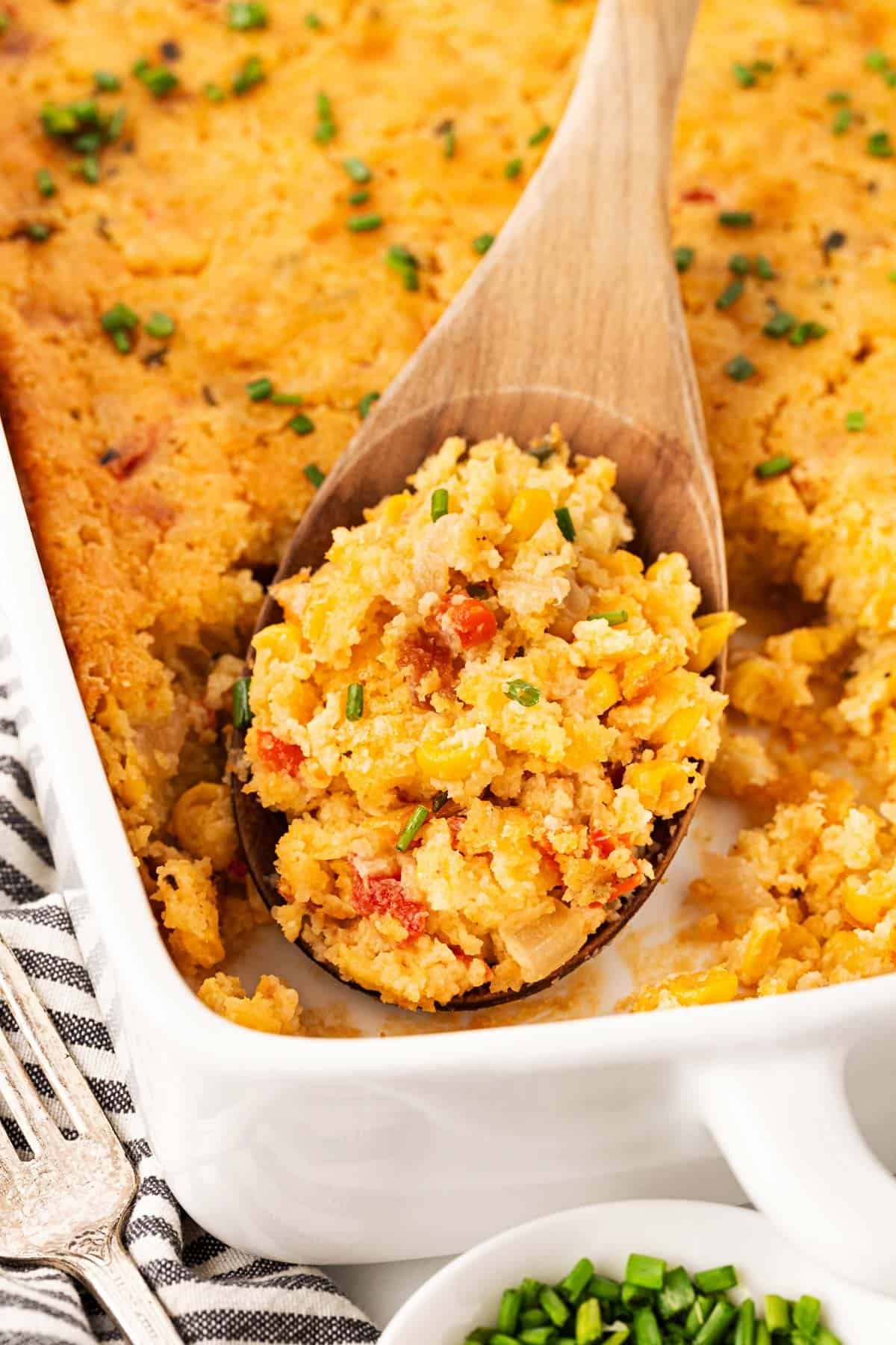 Closeup of a wooden spoon scooping a portion of baked cornbread pudding from a casserole dish