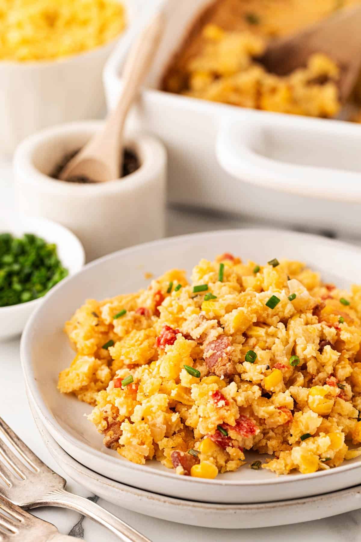 Plate of cornbread pudding garnished with chopped chives, with the baking dish and ingredients blurred in the background