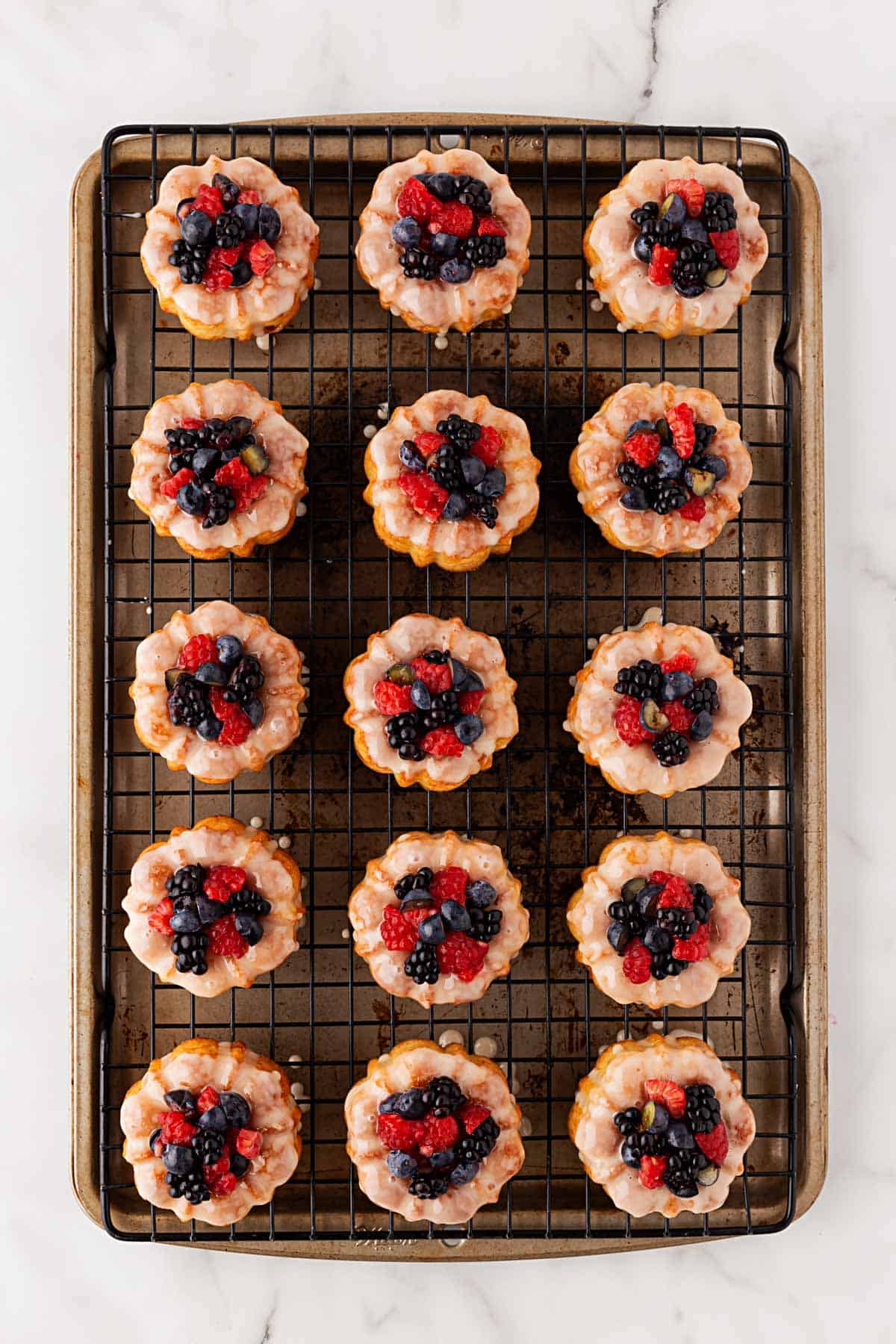 Overhead shot of mini honey bundt cakes on a wire rack, each topped with a light glaze and a mix of fresh berries including raspberries, blackberries, and blueberries