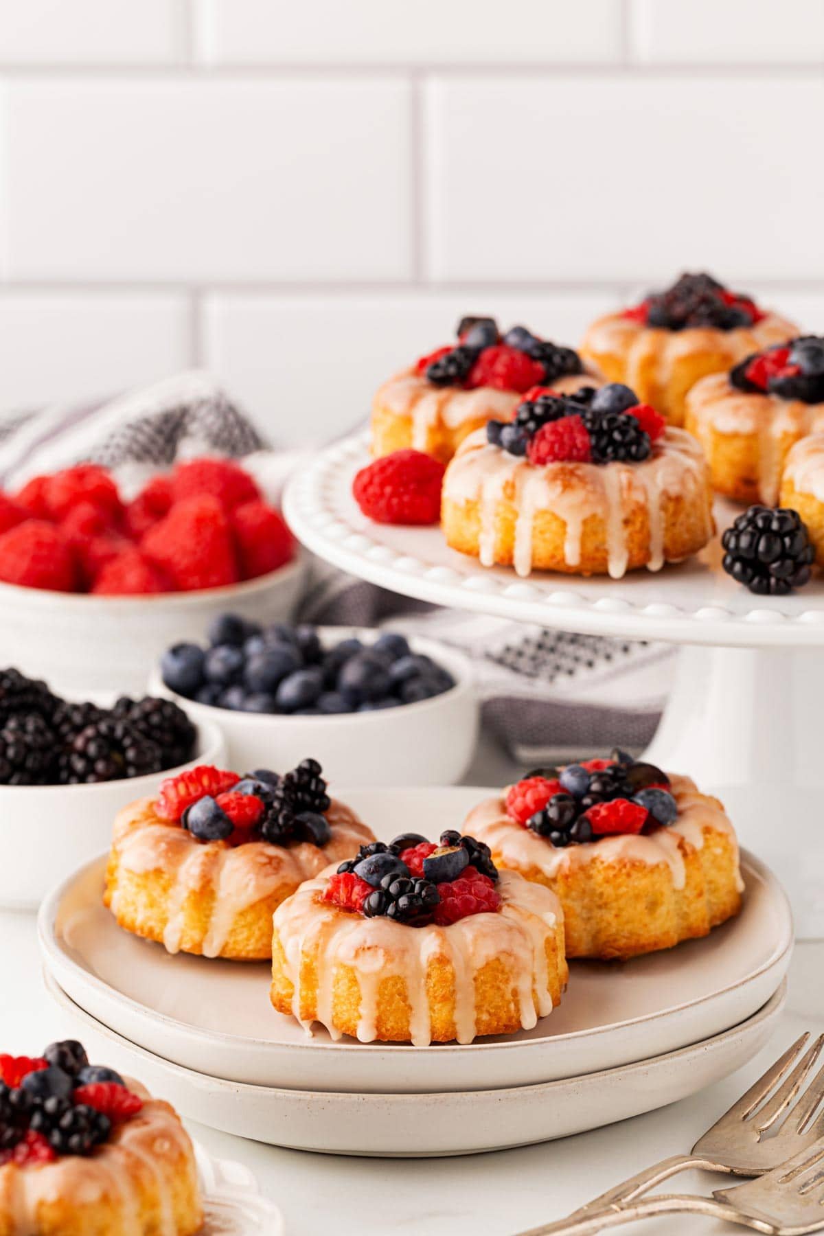 Several mini honey bundt cakes arranged on two white serving stands, with bowls of berries in the background
